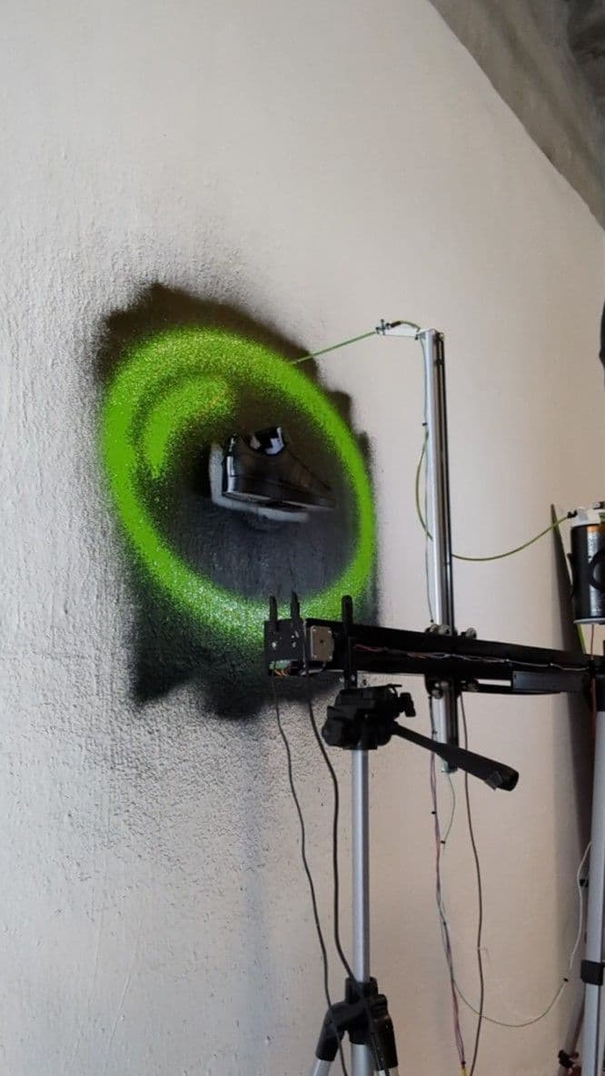 The robotic sprayer is painting illustrations onto an Adidas Superstar shoe that is mounted to the wall.